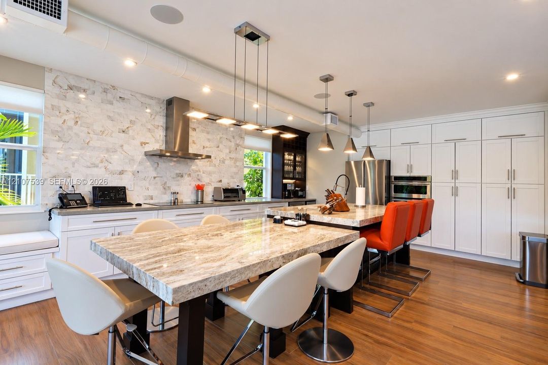 Kitchen featuring stone-topped islands, a floor-to-ceiling cabinetry wall, a natural stone tile backsplash, stainless steel appliances, and wood-finish flooring
