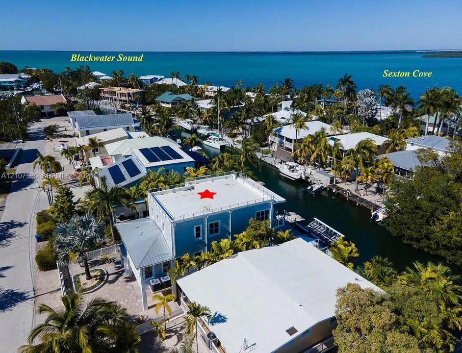 Aerial perspective featuring a multi-level residence with a rooftop deck, metal roof, paved driveway, and canal-front bulkhead