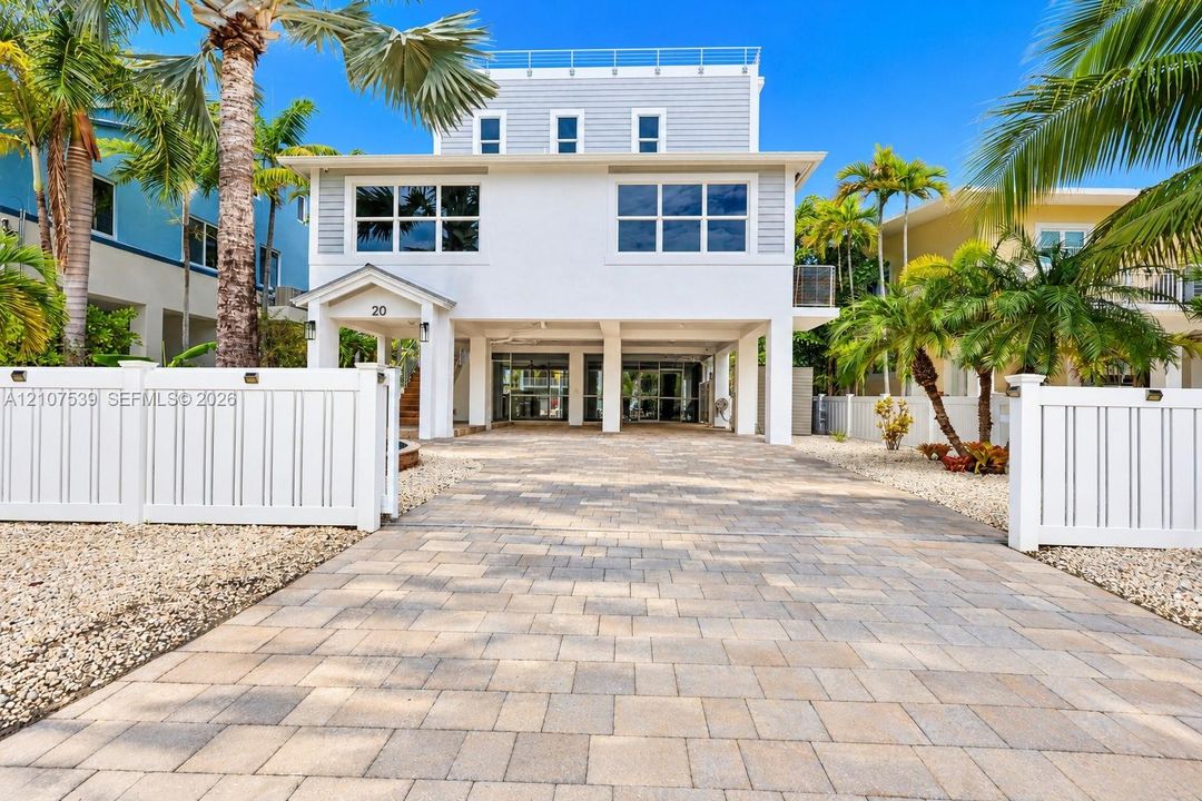 Three-story elevated residence featuring a paver driveway, covered ground-level parking, multi-pane windows, and a white vinyl perimeter fence