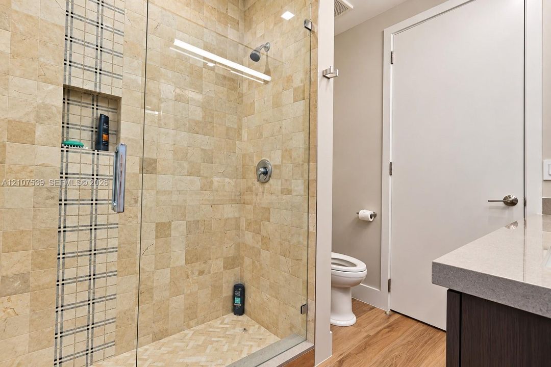 Walk-in shower with square stone tiling, built-in vertical mosaic accent niche, frameless glass door, and a solid-surface vanity countertop