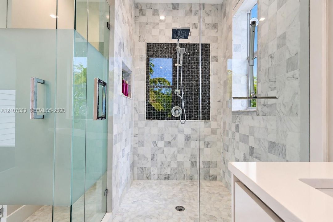 Walk-in shower featuring glass paneling, floor-to-ceiling stone-tiled walls, a mosaic accent inlay, a wall-mounted shower system, and a built-in recessed shelving niche