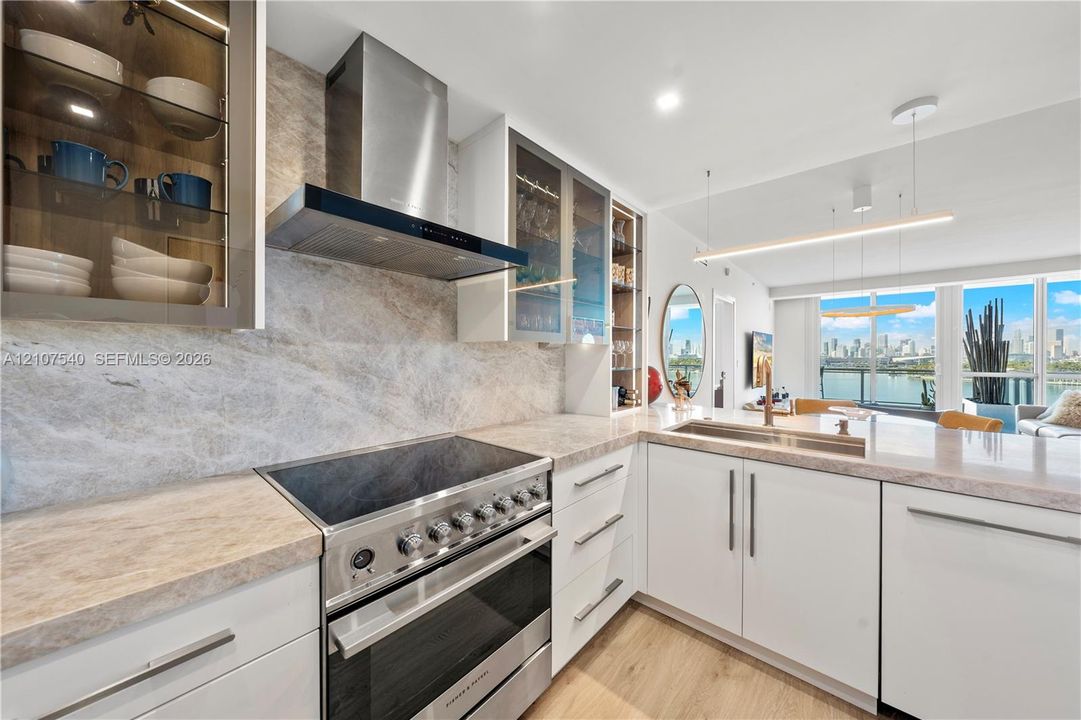 Gourmet Kitchen featuring Taj Mahal Quartzite countertops, full-height Taj Mahal stone backsplash, glass-front cabinetry with interior lighting, Fisher & Paykel gourmet appliances with overhead vent hood, and LED pendant lighting