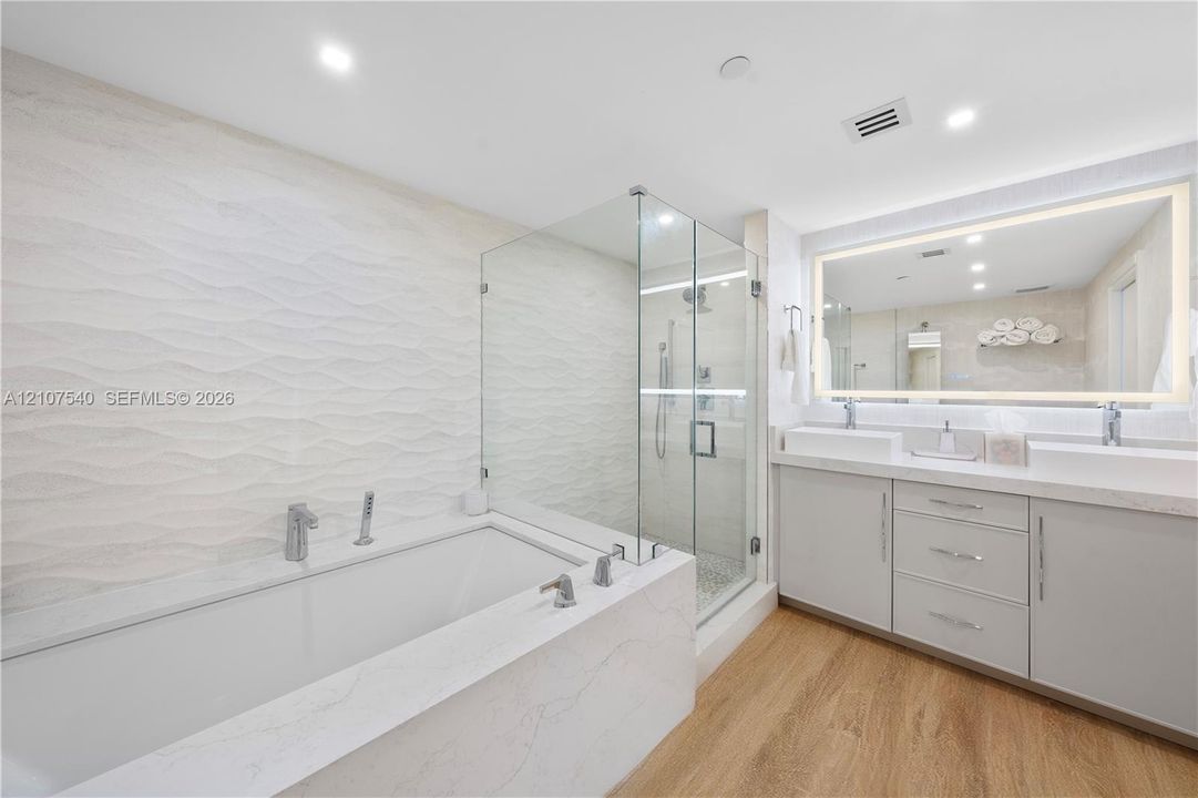 Master bathroom featuring a frameless glass-enclosed shower, a deep soaking tub with stone-finish surround, a dual-sink vanity with comfort height cabinetry, and an oversized back illuminated wall mirror