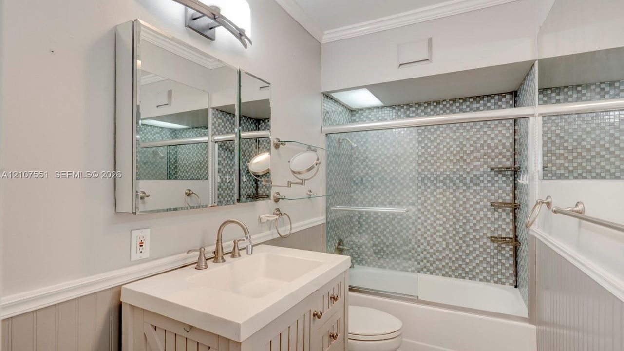 Vanity featuring a rectangular basin, metallic faucet, and integrated storage, paired with a tri-view mirrored medicine cabinet, beadboard wainscoting, and a bathtub with mosaic tile surround and sliding glass doors