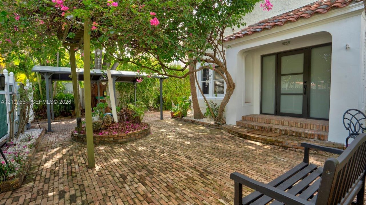 Courtyard entry featuring a herringbone-patterned brick patio, white stucco exterior, terracotta-tiled roof accents, a covered parking structure, and a glass-paneled entry door