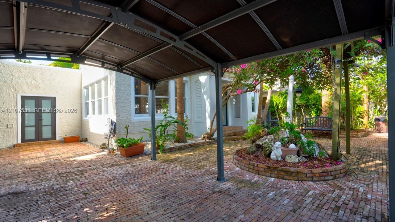 Covered outdoor structure with dark metal frame, brick paver ground surface, and circular brick-bordered landscaping bed