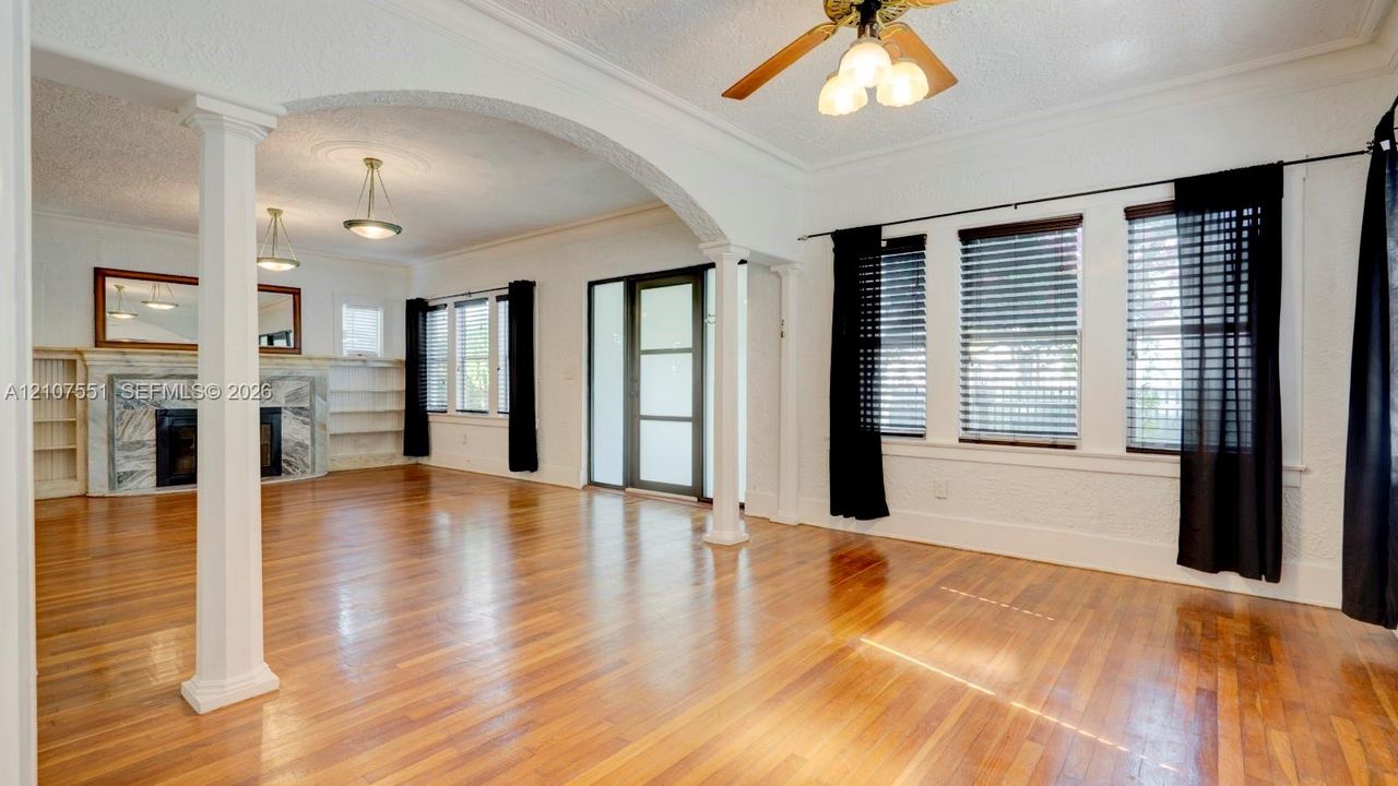 Open living area featuring wood-finish flooring, a stone-surround fireplace, built-in shelving, decorative columns, and an arched architectural opening