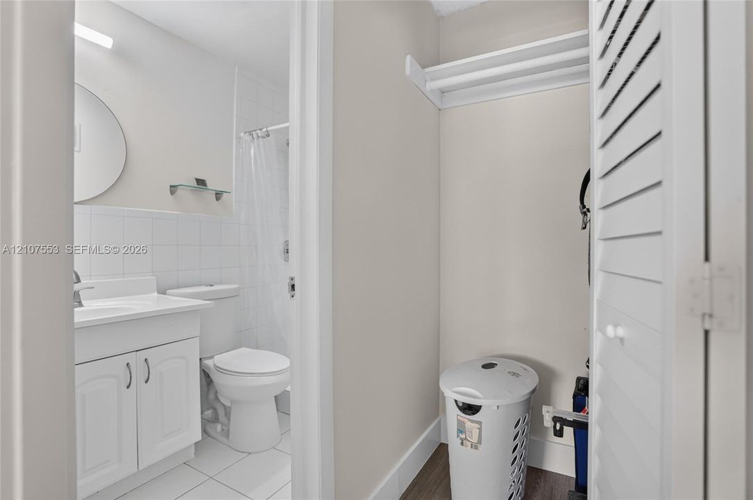 Bathroom featuring a white vanity cabinet, tiled shower surround, wall-mounted circular mirror, and louvered closet doors with integrated shelving