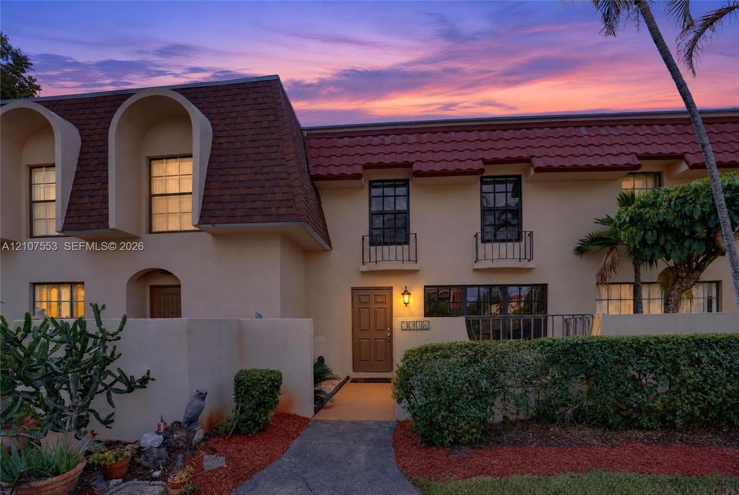 Stucco exterior featuring dual-pitched rooflines, decorative arched dormers, ornamental metal window railings, and an entry courtyard with low-maintenance landscaping