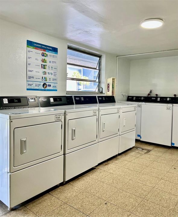 Utility room featuring multiple high-capacity white washing machines and dryers, ceramic tile flooring, a horizontal jalousie window, and overhead flush-mount lighting