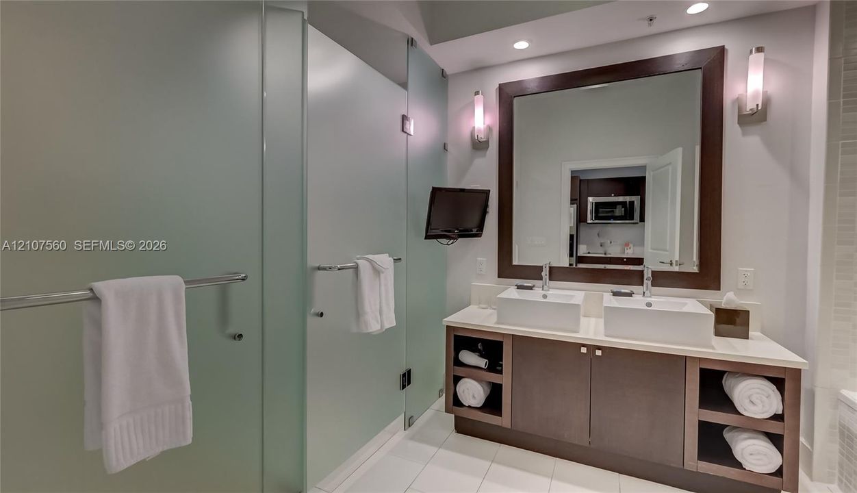 Double vanity featuring rectangular vessel sinks, light-toned stone-surface countertop, dark wood-finish cabinetry with open shelving, large wall-mounted mirror with wood frame, and frosted glass shower enclosure