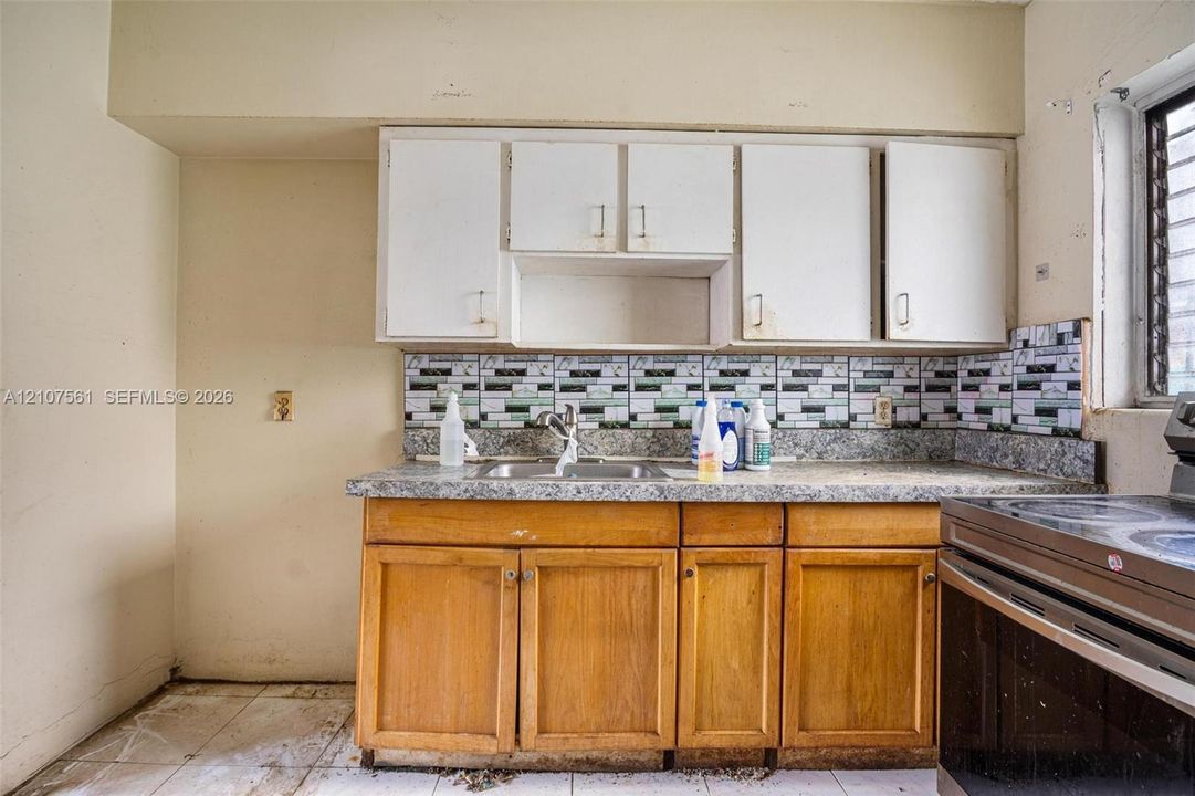 Kitchen featuring wooden lower cabinetry, white upper cabinets, stone-composite countertops, multi-toned decorative tile backsplash, and stainless steel dual-basin sink