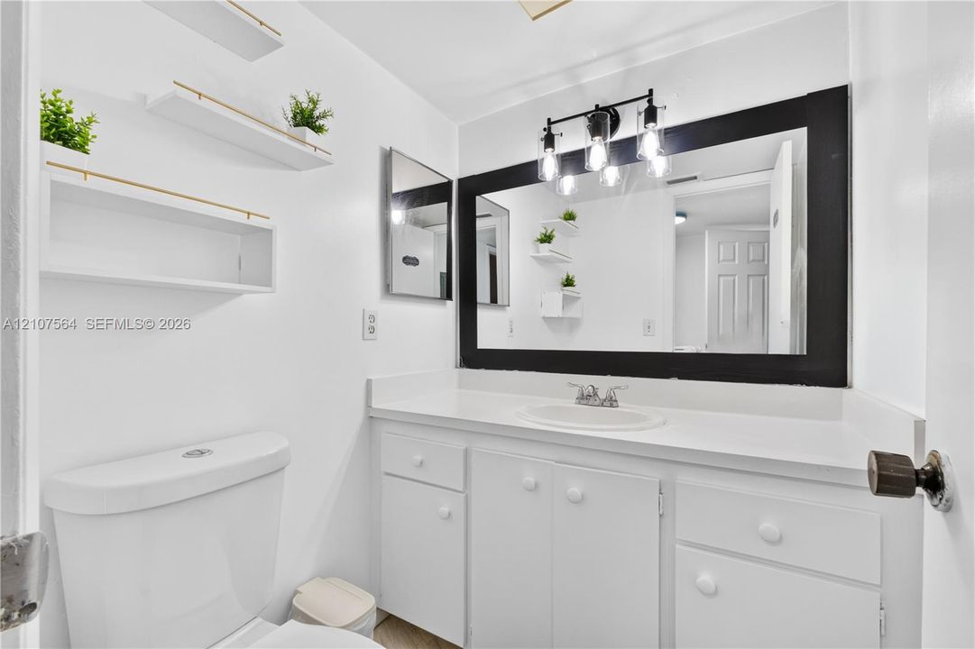 Bathroom featuring a vanity with a white countertop, inset sink, three-light fixture, large black-framed mirror, and wall-mounted white shelving