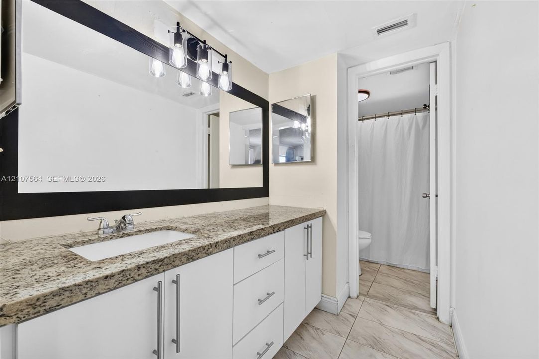 White cabinetry with stainless steel pulls, speckled stone countertop, large rectangular mirror with a dark frame, four-light vanity fixture, and tile flooring