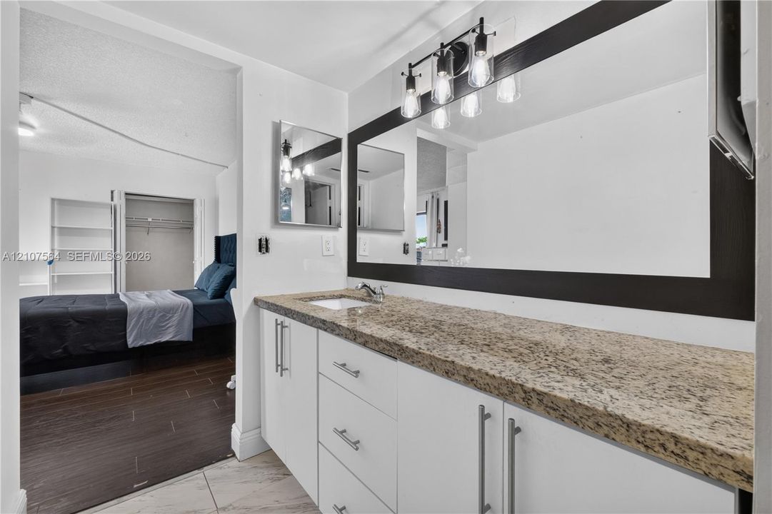 Bathroom vanity featuring a stone-look countertop, integrated sink, white cabinetry with silver-tone hardware, a large framed mirror, and a multi-bulb light fixture