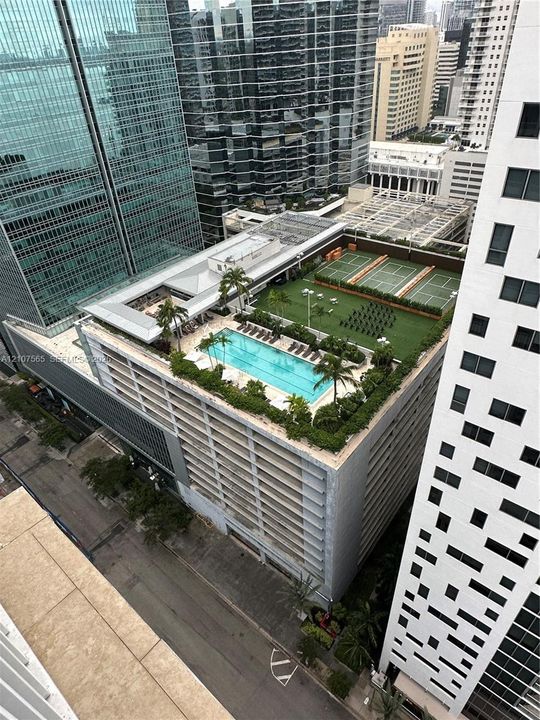 Rooftop amenity deck featuring a rectangular swimming pool, tennis courts, synthetic turf lawn area, and perimeter landscaping
