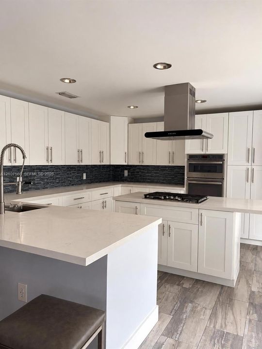Kitchen featuring white shaker-style cabinetry, solid-surface countertops, a dark mosaic tile backsplash, stainless steel built-in double wall ovens with a gas cooktop, and wood-finish flooring