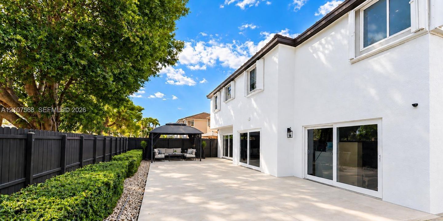 Exterior rear elevation featuring white stucco siding, dual sliding glass doors, a stone-paver patio, and perimeter privacy fencing accented by manicured hedges