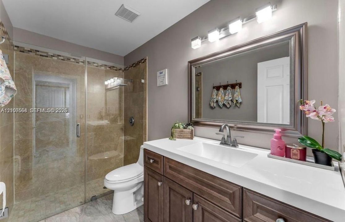 Bathroom featuring a wood-finish vanity with a rectangular sink and solid-surface countertop, a framed wall mirror, four-light vanity fixture, and a glass-enclosed shower with tiled surround