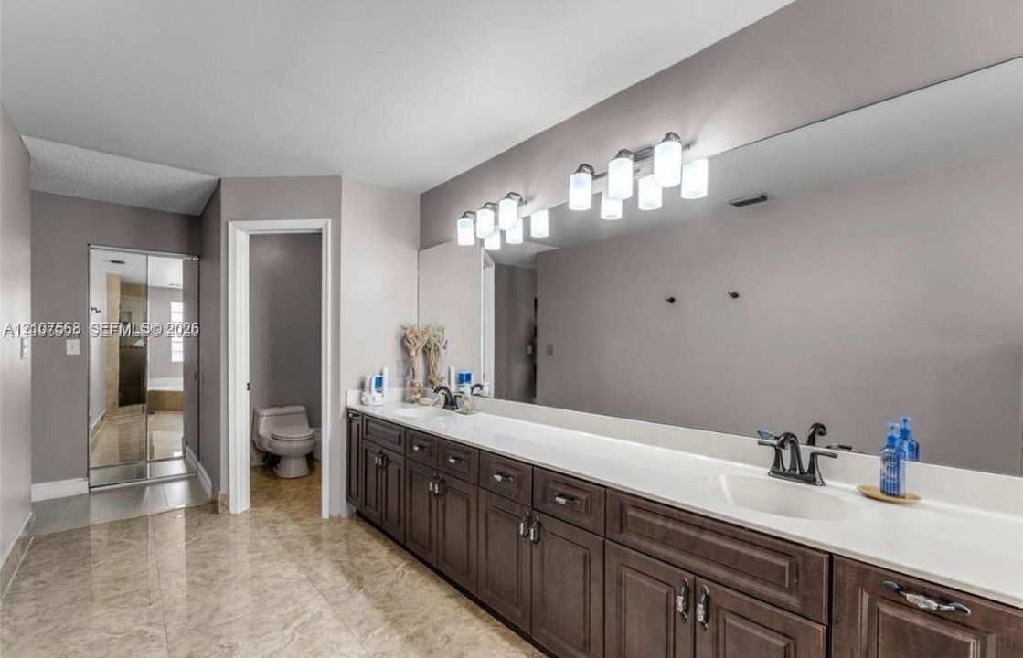 Extended vanity with dual undermount sinks, stone-finish countertop, dark wood cabinetry, expansive wall-to-wall mirror, and multi-bulb vanity lighting