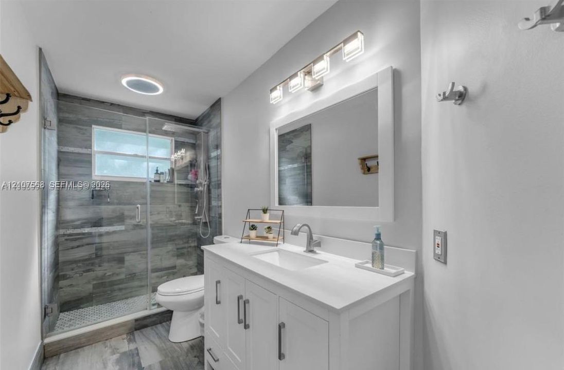 White vanity with integrated basin and polished chrome faucet, glass-enclosed shower featuring wood-finish tile surround, rectangular vanity mirror, and linear wall-mounted lighting