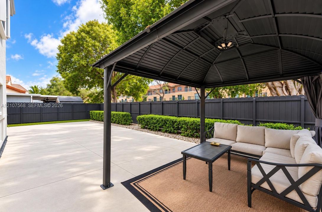 Concrete patio featuring a permanent metal gazebo with integrated lighting, enclosed by a privacy fence and manicured hedge border