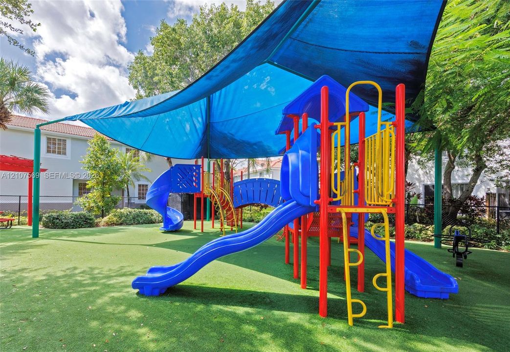 Outdoor recreational area featuring a multi-level playground structure, synthetic turf ground cover, and a large overhead shade canopy
