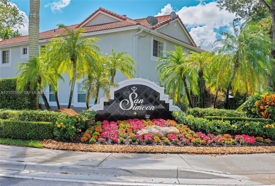 Stucco exterior with a tile roof, complemented by a professional monument sign and manicured landscaping featuring flowering shrubs and palm trees