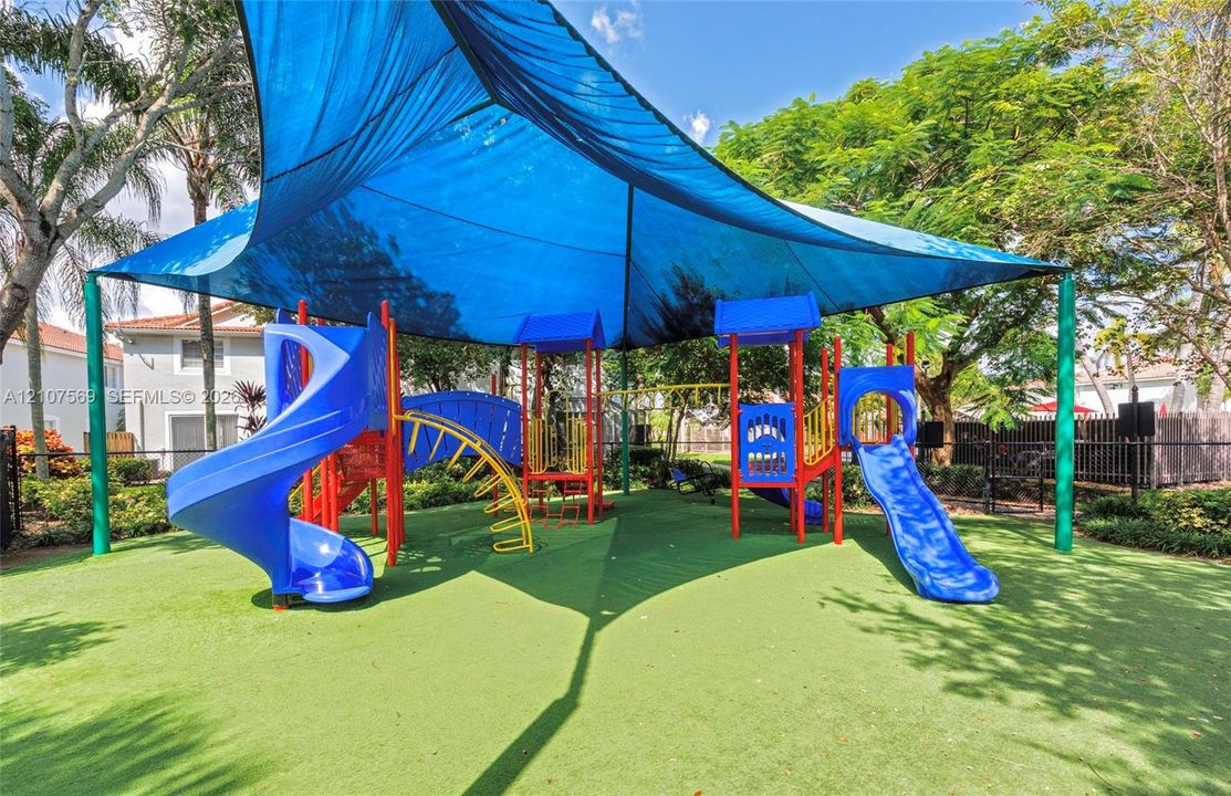 Recreational area featuring a multi-component play structure with dual slides and overhead shade canopy set atop synthetic turf