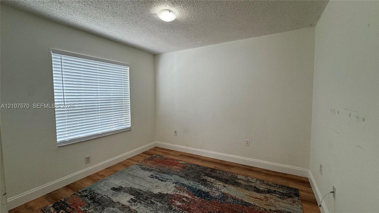 Neutral-toned interior room featuring wood-finish flooring, a single window with horizontal blinds, and a central ceiling-mounted light fixture
