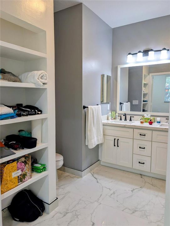 Bathroom featuring white vanity cabinetry with matte black hardware, dual-sink stone-finish countertop, full-width framed mirror with four-light vanity fixture, white tile flooring with veining detail, and built-in floor-to-ceiling shelving