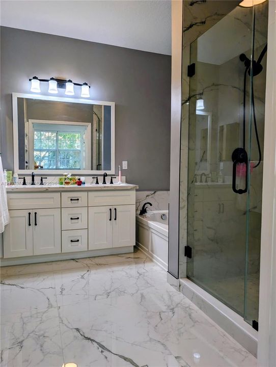 Double vanity with white cabinetry, stone-patterned countertop, glass-enclosed shower, integrated soaking tub, and large-format veined flooring
