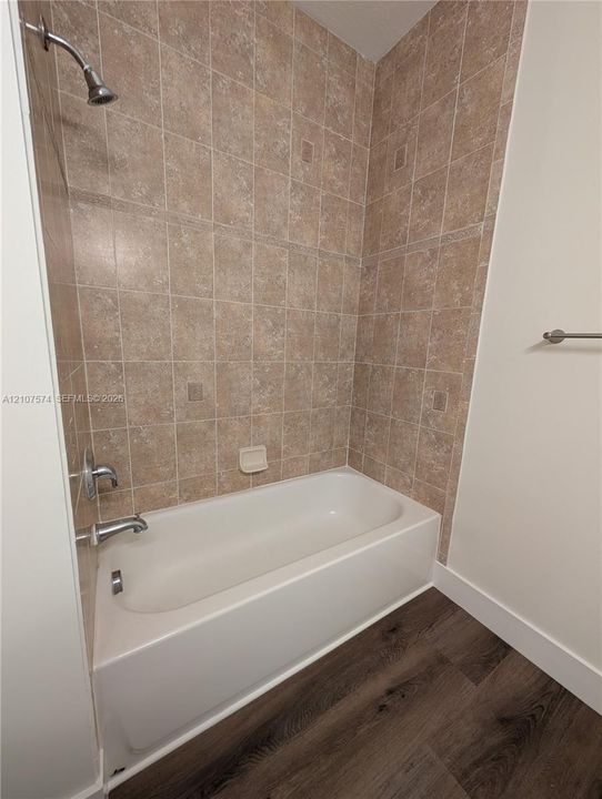 Bathtub with a showerhead, beige square tile surround, built-in soap dish, chrome fixtures, and wood-finish flooring