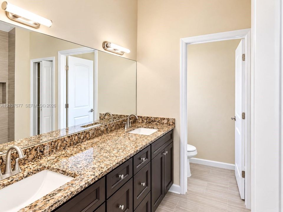 Dual-sink vanity with stone-finish countertops, dark cabinetry, large wall-mounted mirror, and tile flooring