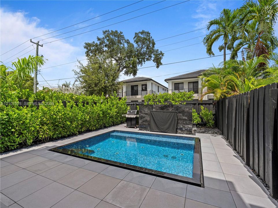 In-ground pool with tile surround, stone-finish accent wall, and perimeter hedge privacy screening
