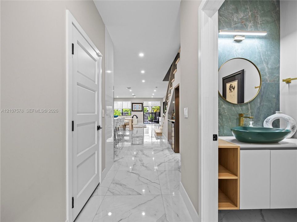Hallway featuring large-format tile flooring and recessed lighting, with a powder room showcasing a green stone slab backsplash, vessel sink, and built-in vanity cabinet