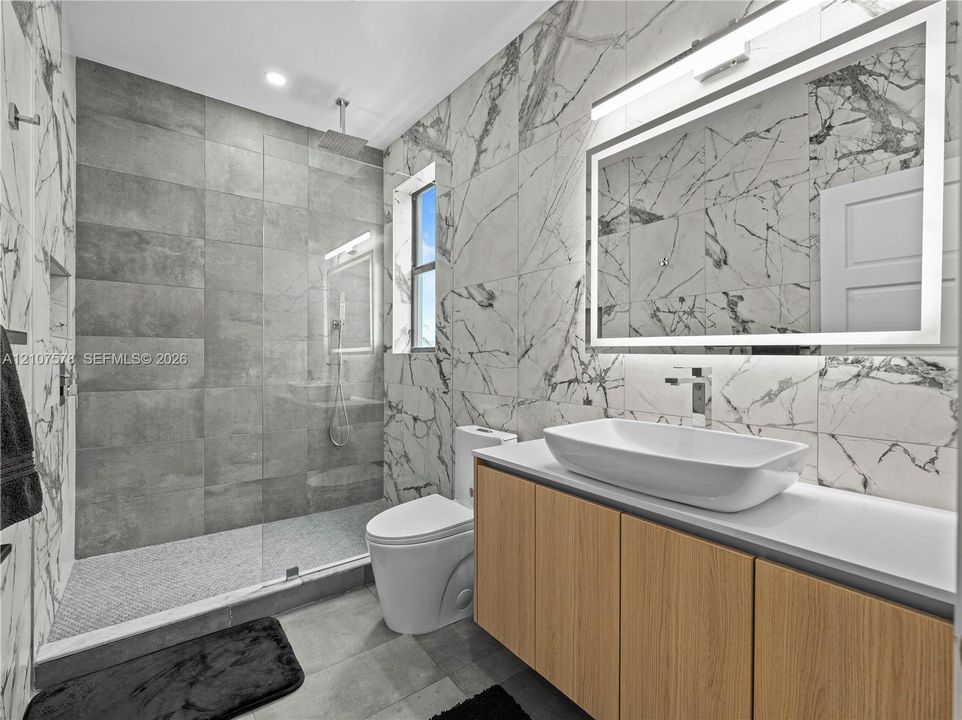 Bathroom featuring large-format wall-to-wall stone-style tile, a walk-in glass shower enclosure with a rainfall showerhead, a wood-finish vanity with a rectangular vessel sink, and a large illuminated vanity mirror