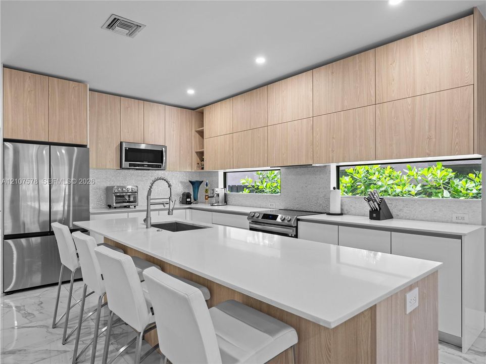 Contemporary kitchen featuring light wood-finish cabinetry, white solid-surface countertops, a center island with integrated sink, stainless steel appliances, and ribbon windows