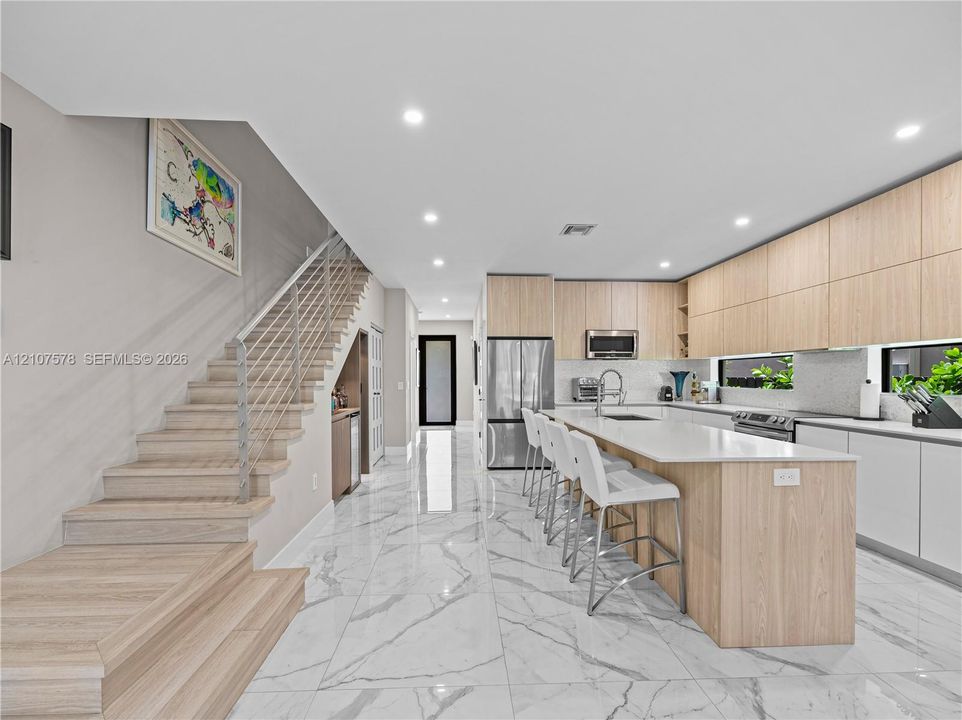 Modern kitchen featuring light wood-finish cabinetry, a large central island with solid-surface countertops, polished stone-finish floor tiling, and a custom staircase with a sleek horizontal railing