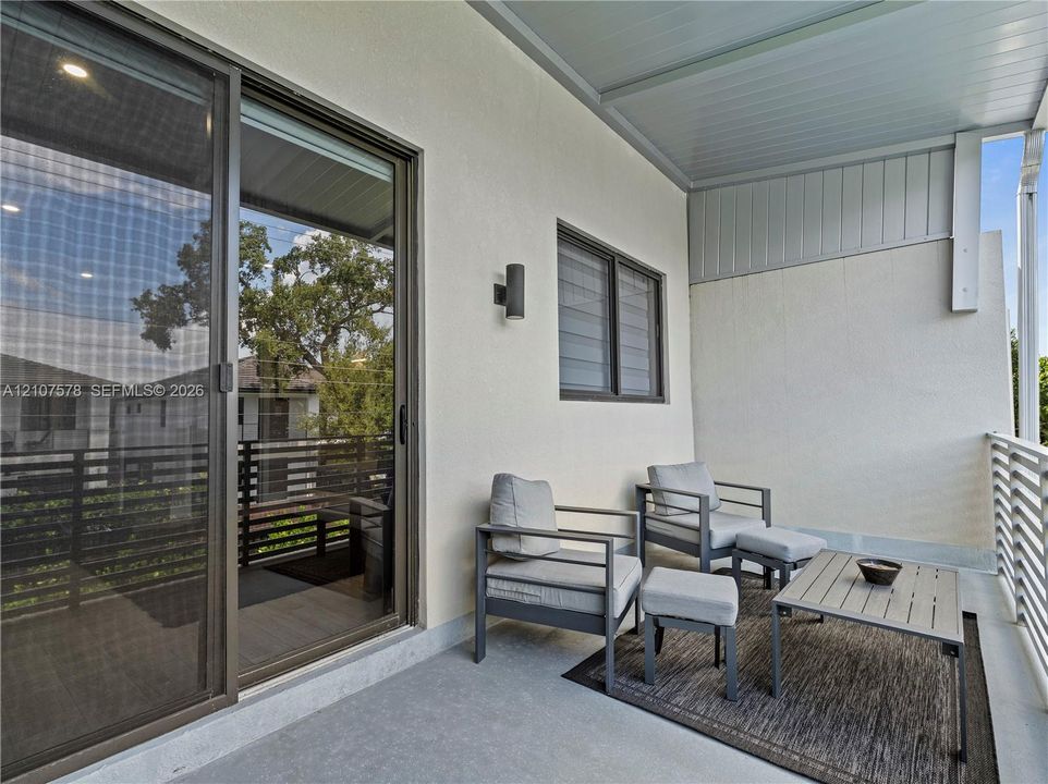 Covered balcony featuring sliding glass doors, horizontal slat railings, neutral stucco exterior wall, and wall-mounted exterior lighting
