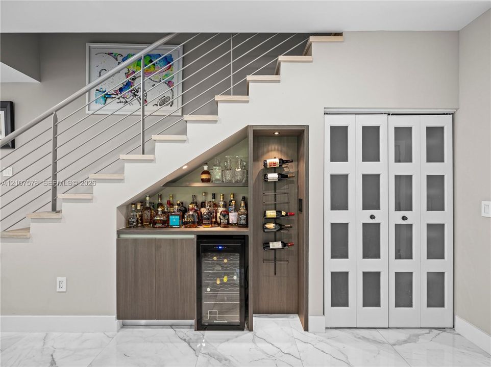 Built-in wet bar featuring wood cabinetry, integrated wine refrigerator, vertical wall-mounted wine rack, and recessed accent lighting, positioned beneath a staircase with metal railing and finished with stone-style tile flooring