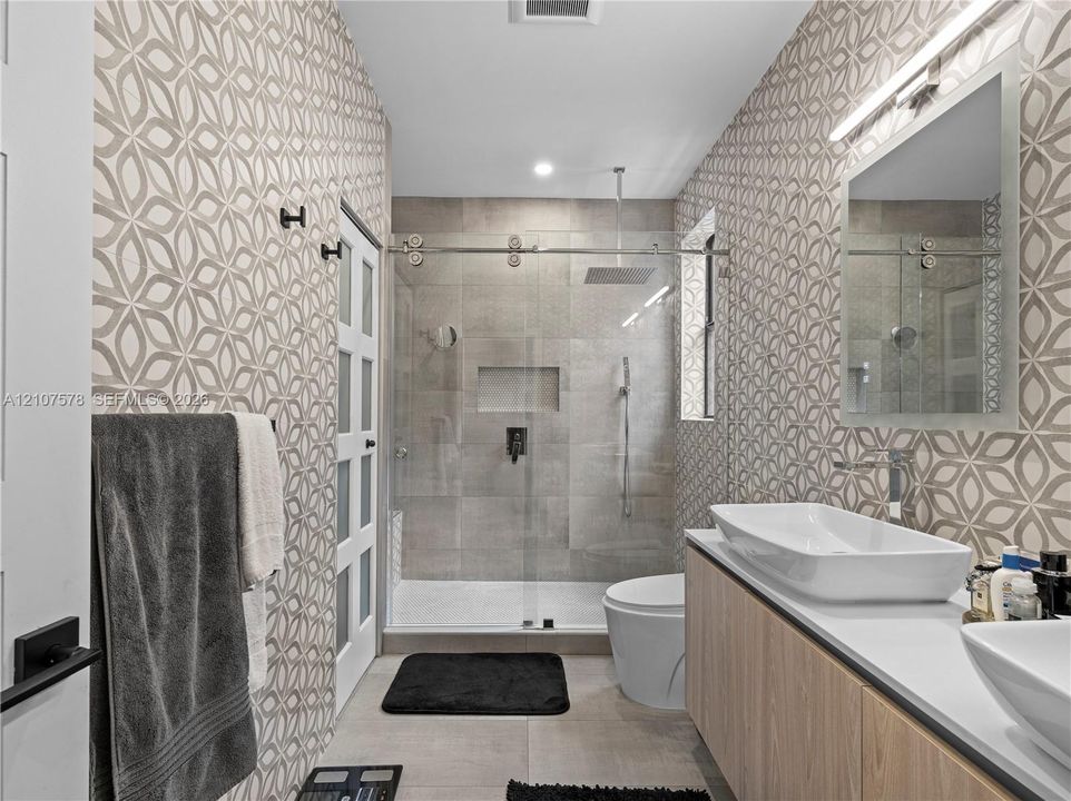 Floating wood-finish vanity with dual vessel sinks, walk-in shower with glass enclosure and rain-style showerhead, patterned wall tile, and built-in recessed shower niche