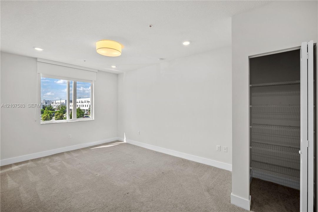 Neutral-toned interior featuring wall-to-wall carpeting, recessed lighting, a central drum light fixture, large window with roller shade, and a closet with white bi-fold doors and built-in wire shelving