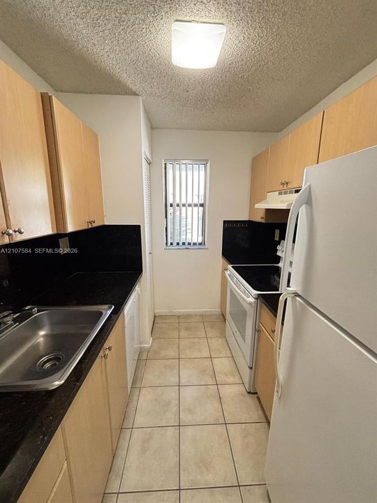 Galley kitchen featuring light-toned cabinetry, dark solid-surface countertops, ceramic tile flooring, and stainless steel sink