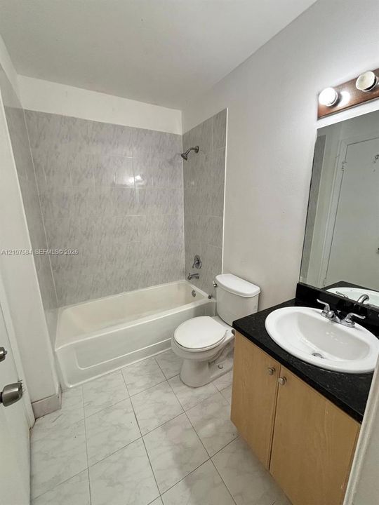 Bathroom featuring a vanity with wood-finish cabinetry, a contrasting dark countertop, an integrated oval sink, and a tiled shower-tub surround with stone-patterned surfacing