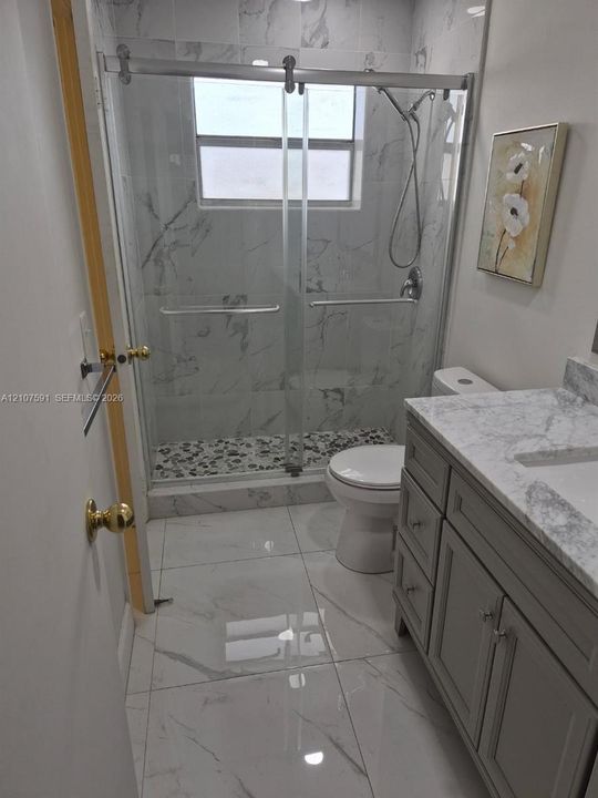 Bathroom featuring a gray vanity with stone-style countertop, shower enclosure with sliding glass doors, stone-style wall and floor tiling, and a built-in pebble-style shower floor