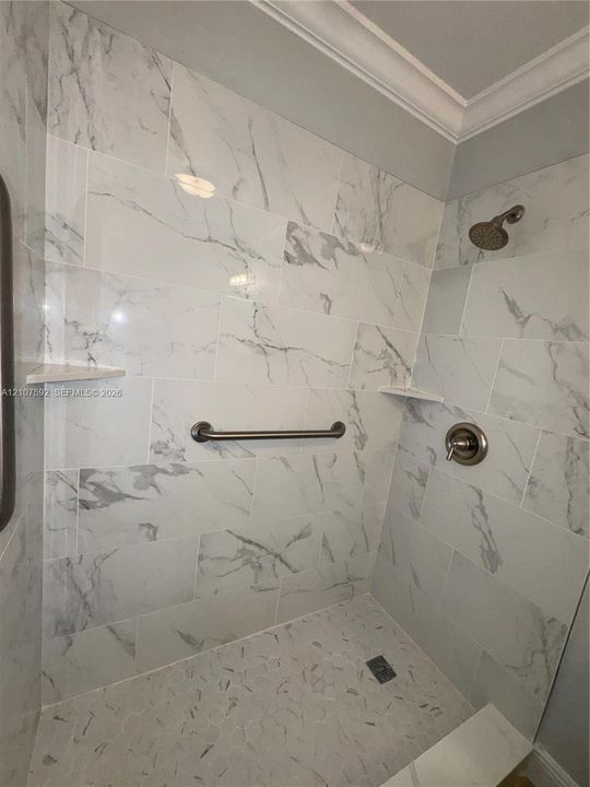 Shower enclosure featuring large-format wall tile with a neutral veined pattern, built-in corner shelving, stone-texture floor tiling, and a metallic finish shower head with matching control handle