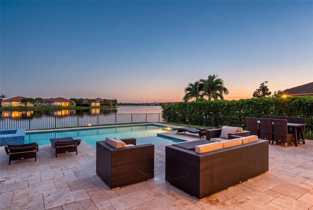 Rear patio featuring travertine pavers, a rectangular swimming pool, a spillover spa, and a metal perimeter fence overlooking the water