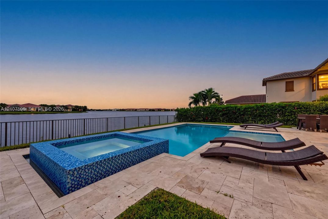 In-ground swimming pool and raised spa with blue tile accents, surrounded by a stone-paver deck, enclosed by a perimeter fence, overlooking a water feature