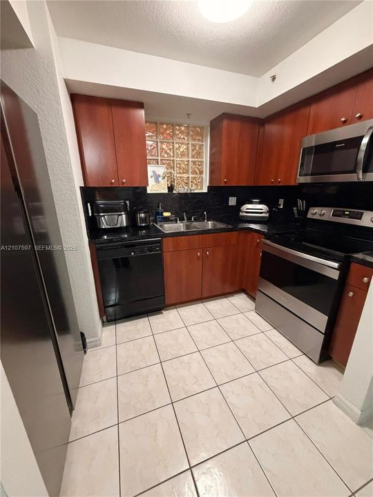 Kitchen featuring wood-finish cabinetry, stainless steel appliances, dark stone-style countertops, ceramic tile flooring, and glass block accent window